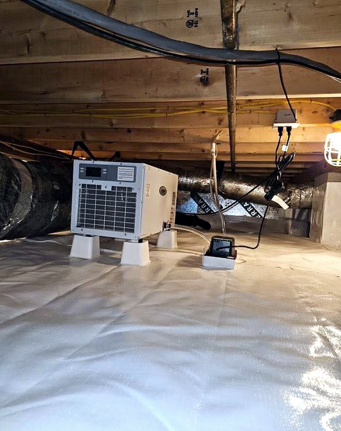 A white crawlspace dehumidifier raised on four stands above a sealed white liner, its drain line running to a small pump, with insulated ductwork behind.
